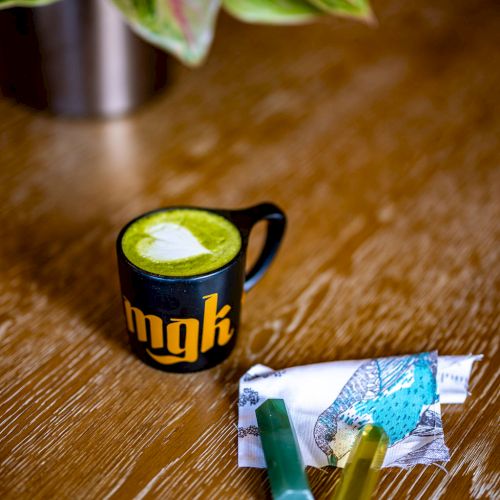 A black mug with yellow text, a green drink, and a package with two colored objects are on a wooden table next to a potted plant.