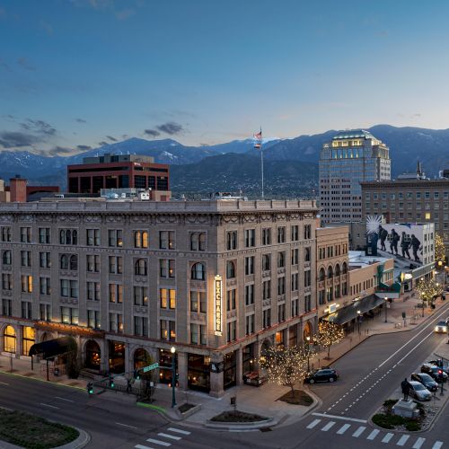 A cityscape at dusk with illuminated buildings, streetlights, cars, mountains in the background, and a clear sky above the urban scene.