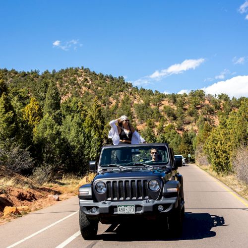 A person sits atop a moving Jeep on a scenic road surrounded by trees and hills under a clear blue sky, with another person driving the Jeep.