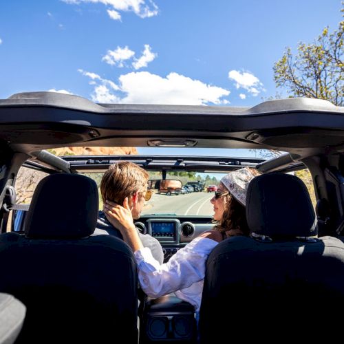 A couple is driving on a scenic road in a convertible car, enjoying a sunny day with a clear blue sky and surrounding nature.