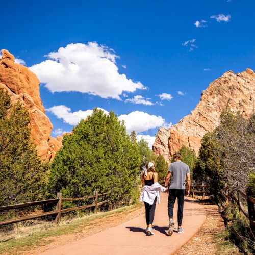 Two people walk along a pathway surrounded by trees and rocky formations under a clear blue sky with clouds.