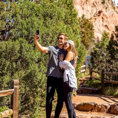 A couple is taking a selfie outdoors on a trail with rocky cliffs and trees in the background, enjoying a scenic and sunny day.
