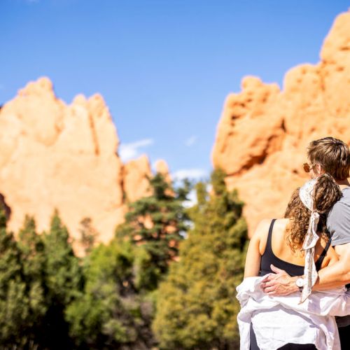 A couple embracing while looking at large rock formations, surrounded by trees and under a clear blue sky.
