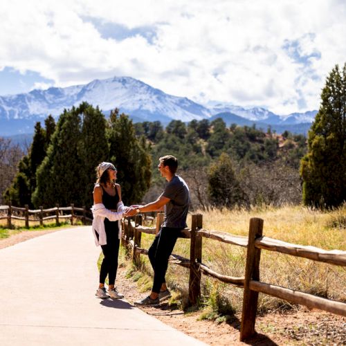 Two people stand on a paved path surrounded by trees and mountains, engaging in conversation under a bright sky.