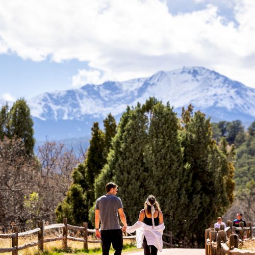 A couple is walking on a paved path surrounded by trees and wooden fences, with a snow-capped mountain in the background under a partly cloudy sky.