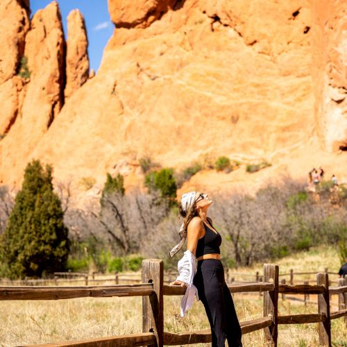 A person stands on a fenced path, gazing up at towering red rock formations under a bright blue sky, with greenery and shrubs in the background.
