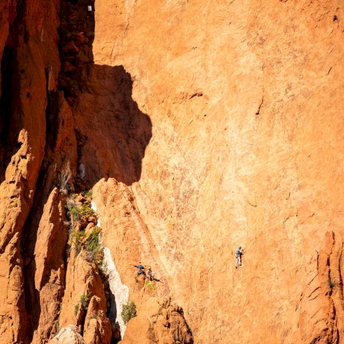 Two rock climbers scaling a steep, towering, reddish-orange cliff with vegetation at the base, amidst a dramatic and rugged landscape.