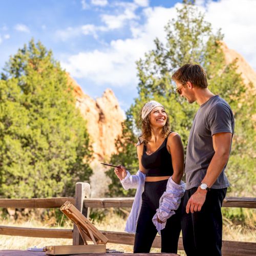 Two people smiling and interacting at a campsite with wooden fencing, with trees and rock formations in the background.