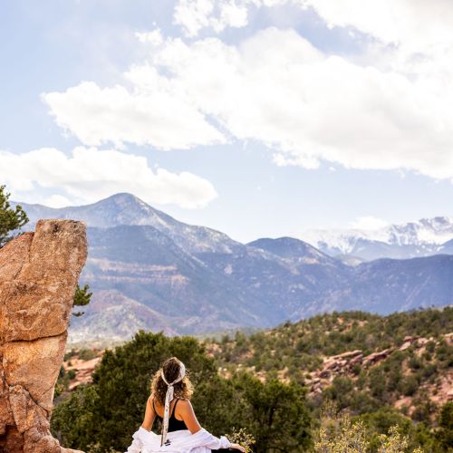 A couple dressed in white sits on rocky terrain, facing a mountainous landscape with clear skies and scattered clouds in the background.
