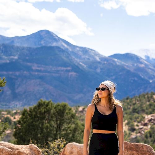 A person wearing sunglasses and a headband stands among rocks with a mountainous landscape in the background on a sunny day.
