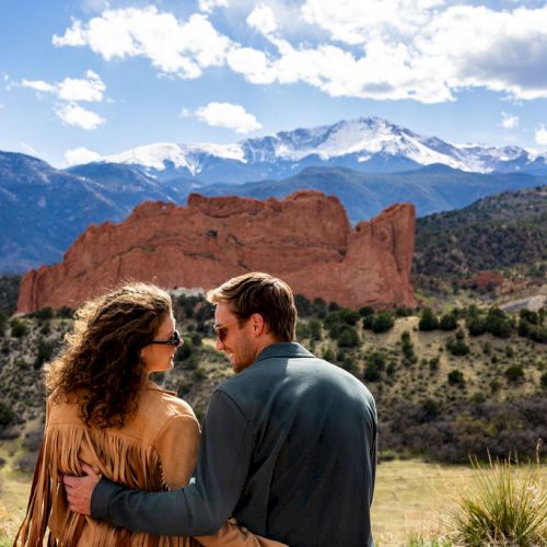 A couple in front of a mountainous landscape with blue sky, clouds, and red rock formations, sharing a tender moment outdoors.