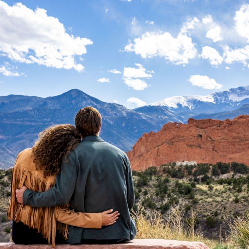 A couple sits on a ledge, embracing and enjoying a scenic view of mountains and red rock formations under a partly cloudy sky.