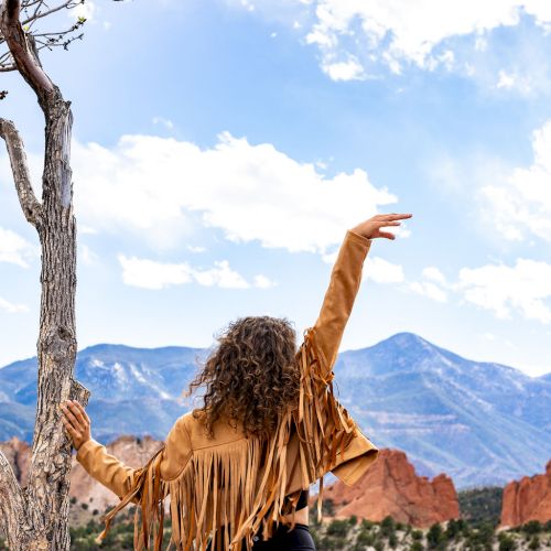A person with curly hair in a fringed jacket raises an arm, standing by a tree, looking at mountains and clouds against a blue sky.