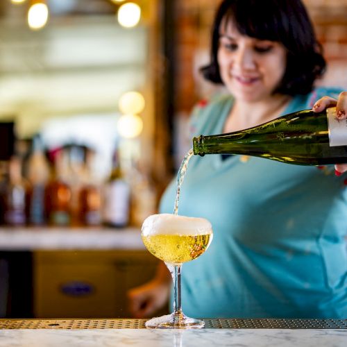 A woman is pouring a drink from a green bottle into a glass at a marble-topped bar, with blurred lights and bottles in the background.