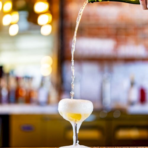 A hand pours sparkling wine or champagne from a bottle into a coupe glass that is beginning to overflow on a bar counter.
