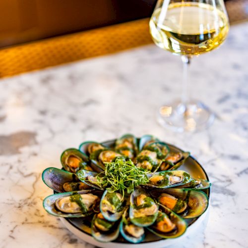 A plate of mussels garnished with greens is placed on a marble table next to a glass of white wine.