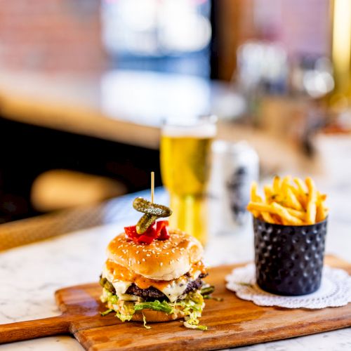 A burger on a wooden board with a side of fries in a metal cup and a glass of beer on a marble counter.