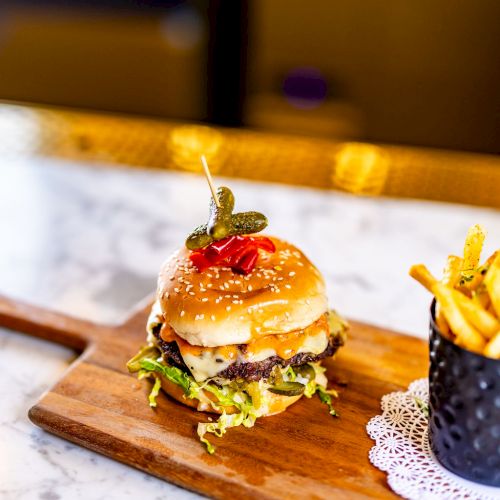 The image shows a burger with various toppings on a wooden board and a small container of fries, garnished elegantly.