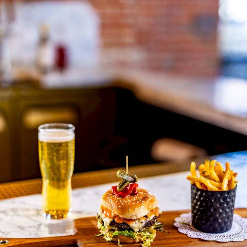 A burger on a wooden board, a container of fries, and a glass of beer are on a marble countertop in a restaurant setting.