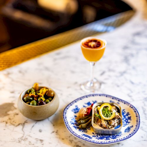 The image shows a marble countertop with a cocktail, a bowl of salad, and a plate of food featuring avocado and garnishes, arranged for a meal.