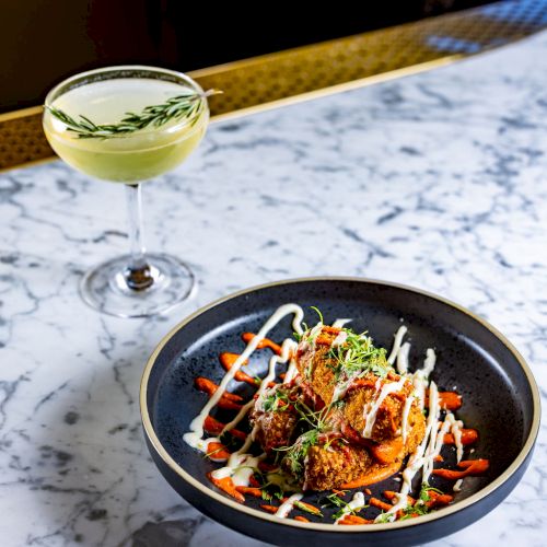 A gourmet dish with sauce drizzled on top, served on a black plate, sits next to a green cocktail garnished with a sprig of herb on a marble table.