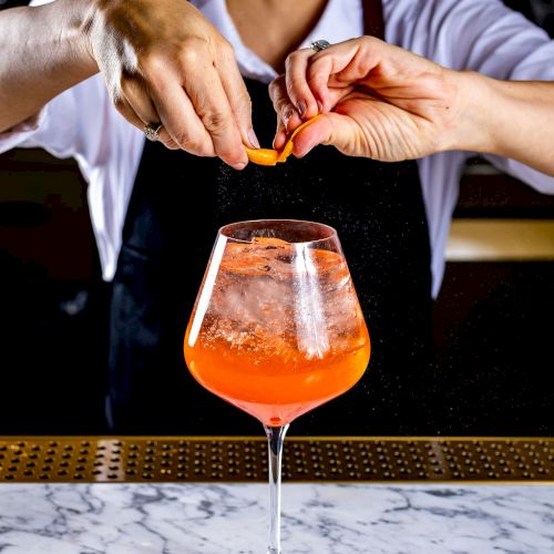 A person is preparing a cocktail by squeezing an orange peel over a glass, smiling. The glass contains an orange drink and is on a marble countertop.