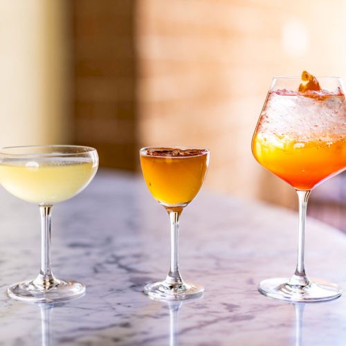 Three cocktails are placed on a marble surface: a yellow drink in a coupe glass, a small brown drink in a shot glass, and an orange drink in a wine glass.