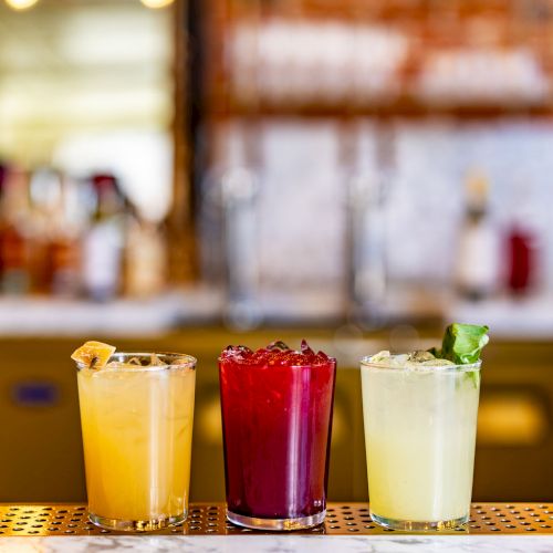 Three colorful drinks on a bar counter: yellow, red, and green, each garnished differently. The background has a blurred bar setting.