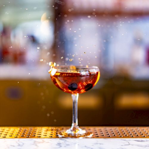 A splash effect captured as liquid is poured into a cocktail glass containing a dark beverage, garnished with a cherry, on a bar counter.