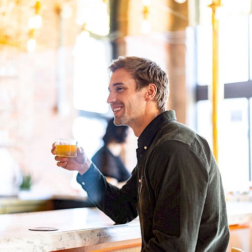 A man sits at a bar holding a glass, smiling, with a blurred background featuring warm lighting and a brick wall.