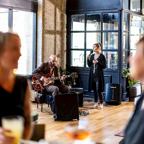 The image shows a cozy indoor setting with a guitarist and a singer performing, while people in the foreground appear to be enjoying their time.