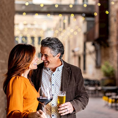 A couple stands close together in an alleyway, holding drinks and smiling at each other, with string lights overhead and an outdoor seating area behind them.