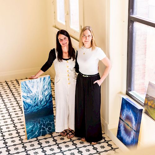 Two women standing in a sunlit room with their artwork displayed, including paintings of nature and abstract scenes, near large windows.