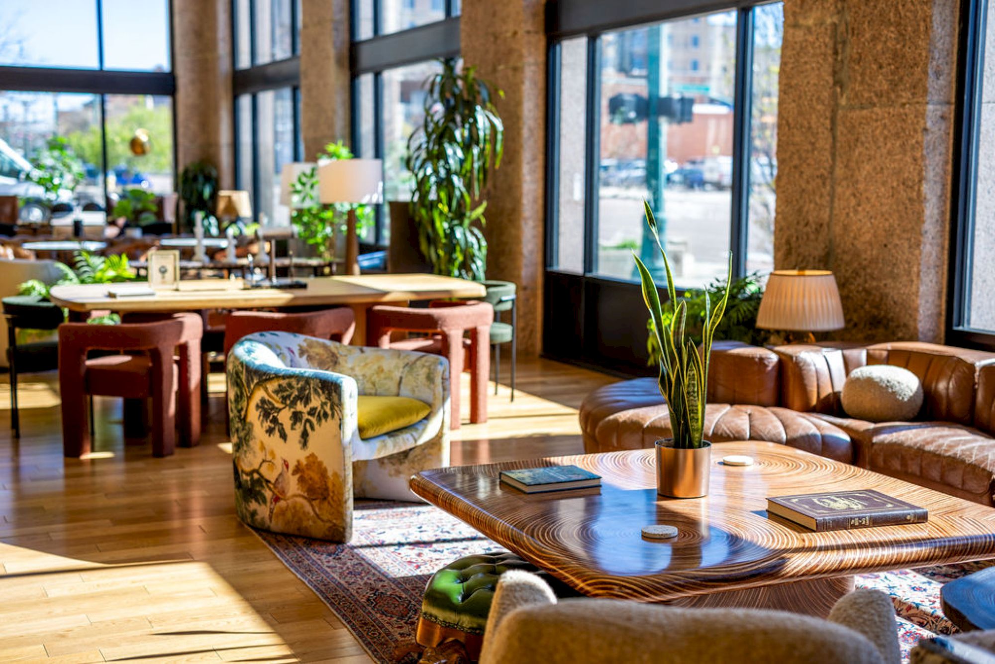 The image shows a well-lit modern lounge with various seating, green plants, tables, and large windows providing a view outside.