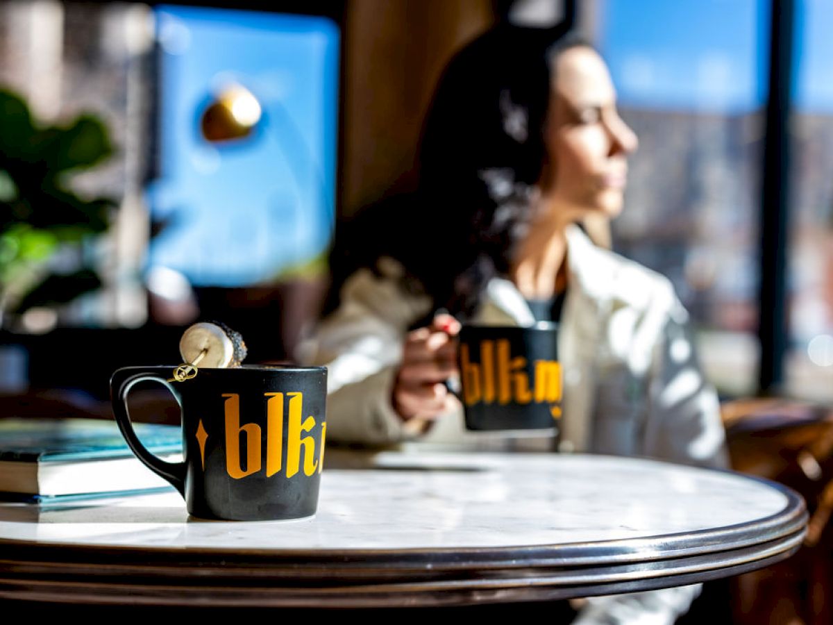 A woman is sitting at a table, holding a black mug with 