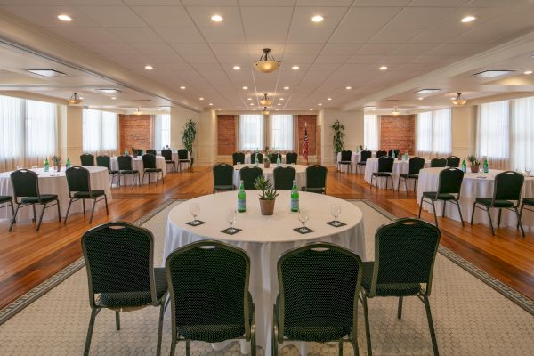 A large conference room with circular tables, each set with chairs, bottles of water, glasses, and notepads. The room has wooden floors and bright lighting.