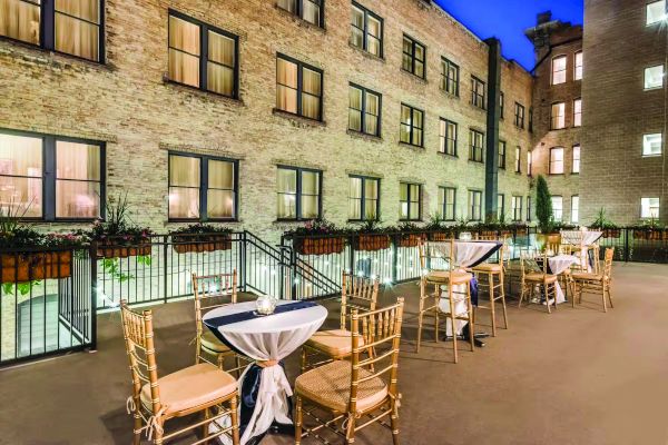 An outdoor patio features several small tables with white tablecloths and wooden chairs. It is surrounded by brick buildings and adorned with plants.