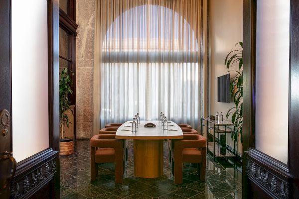A conference room with a long table, six chairs, water bottles, and a TV, flanked by tall curtains, potted plants, and decorative wood panels ends the sentence.