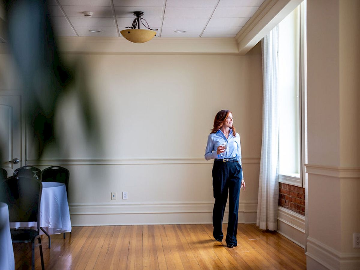 A person is standing by a window in a room with wooden floors and white walls, holding a cup and looking outside.