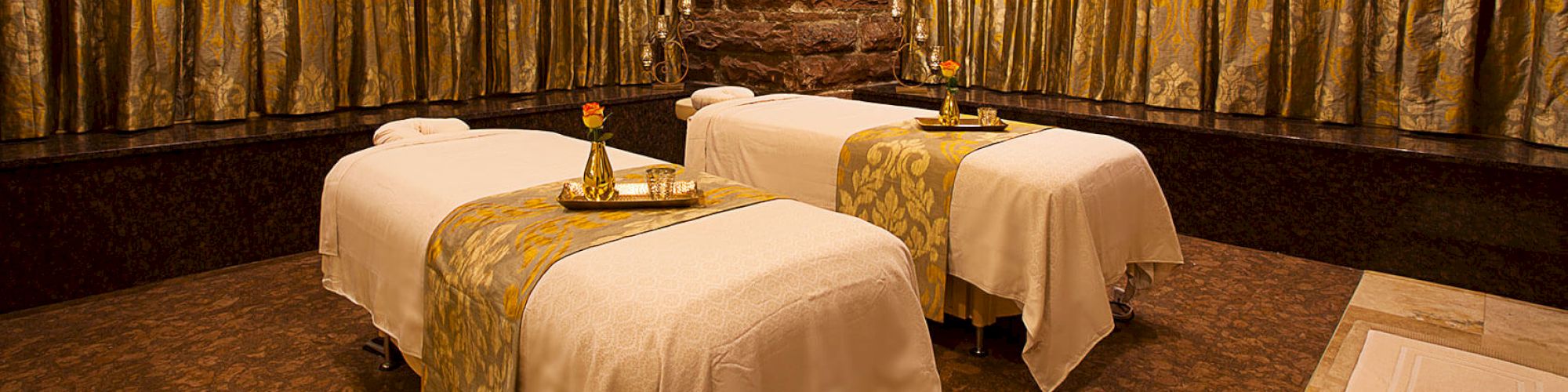 A luxurious spa room with two massage tables covered in white sheets and golden runners, adorned with flowers and a chandelier overhead.