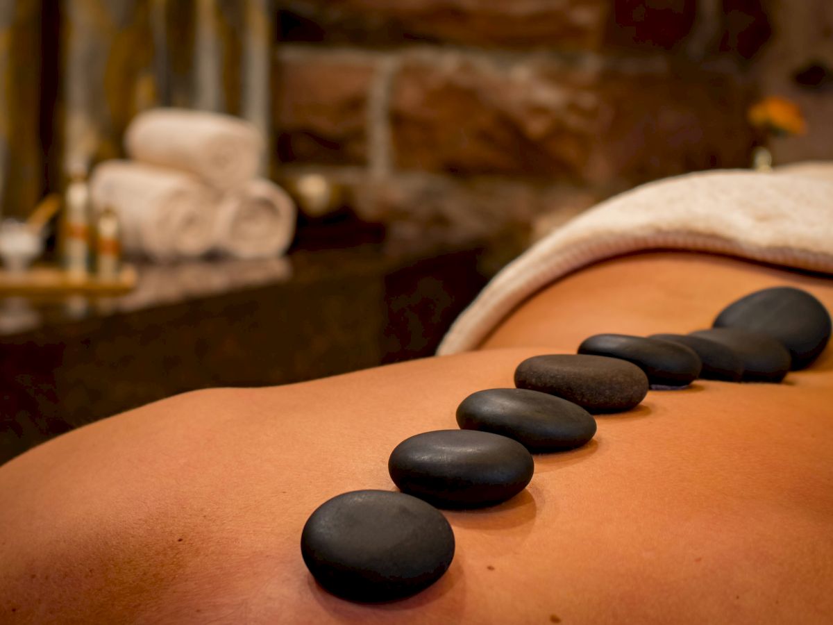 Smooth black stones placed along a person's back in a spa setting, with towels and aromatic oils in the background.