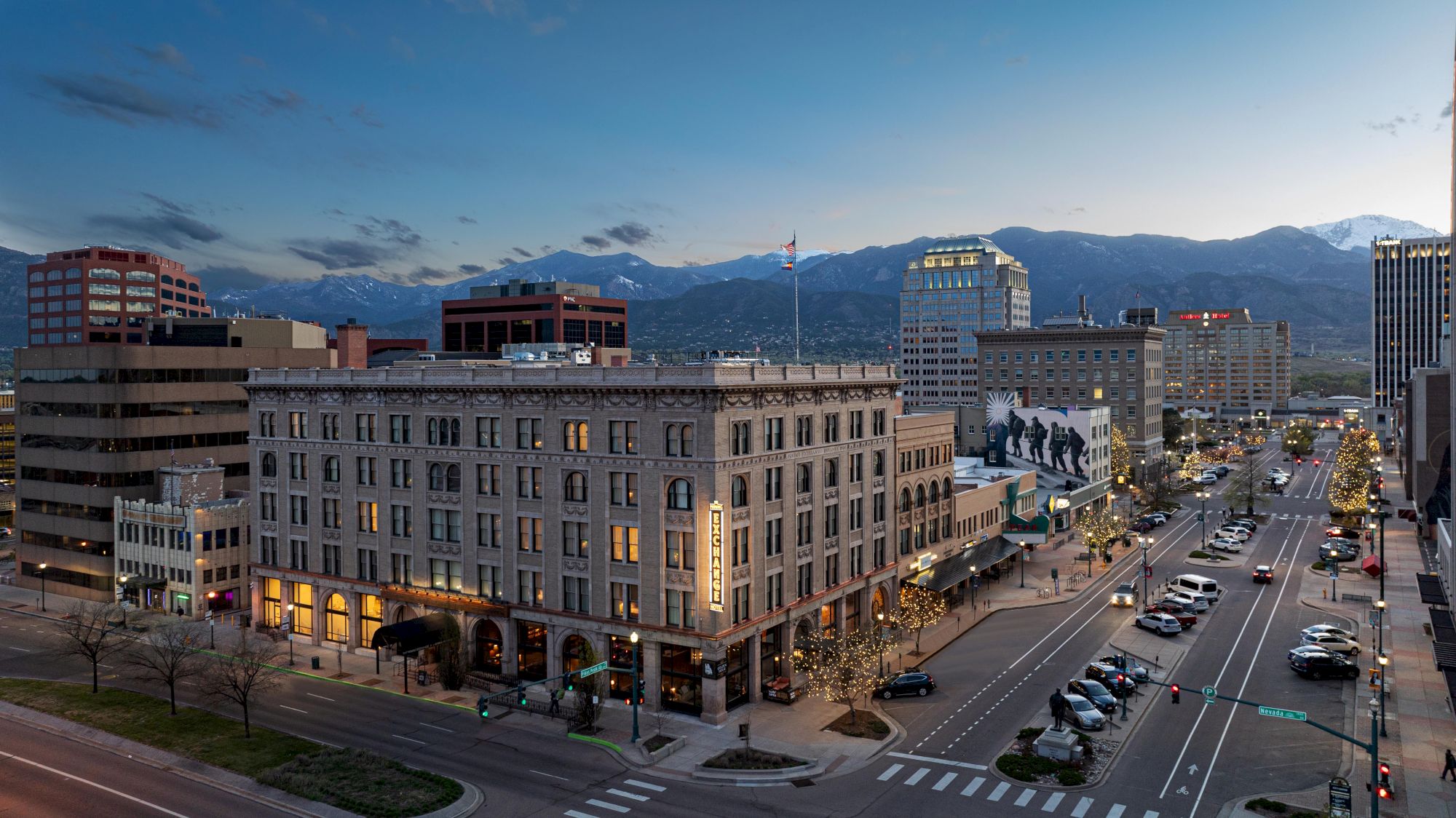 The image shows a city's downtown area at dusk with buildings, a street lined with cars, and mountains in the background.