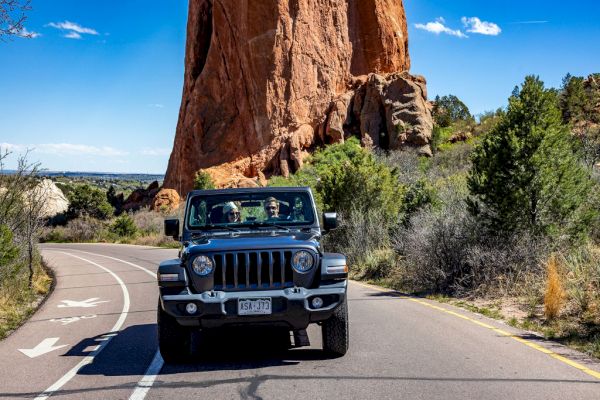 The image shows a Jeep driving on a scenic road with a large rock formation and greenery in the background under a clear blue sky.