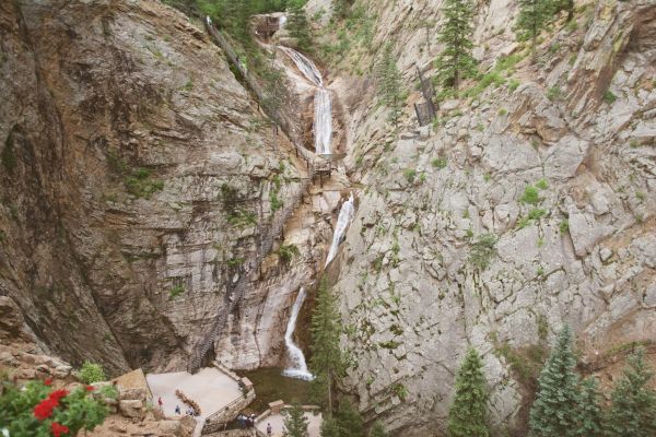 A waterfall cascades through a rocky canyon surrounded by trees, with people gathered at the base on a viewing platform.