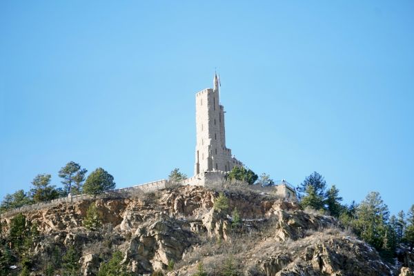 A tall, narrow tower sits atop a rocky hill surrounded by sparse trees under a clear blue sky.
