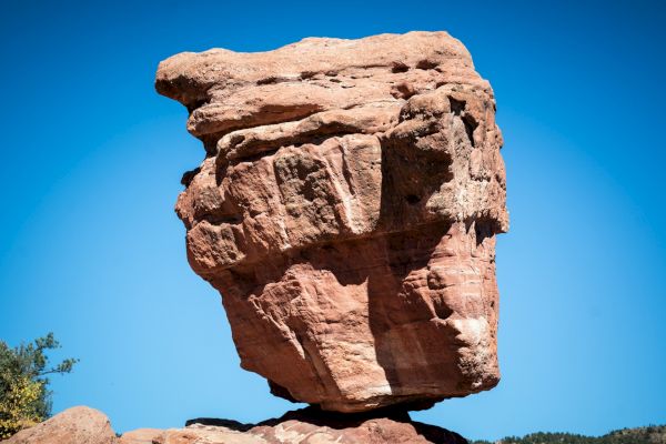 The image shows a massive, balancing rock formation against a clear blue sky. The rock appears to defy gravity, resting on a smaller base.