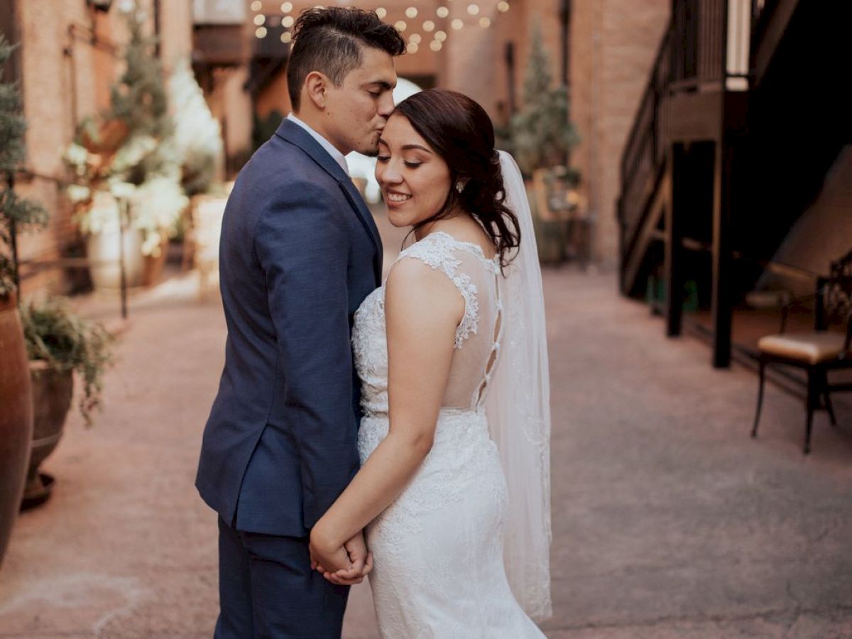 A bride and groom share a tender moment holding hands in a charming, outdoor urban setting adorned with string lights above.