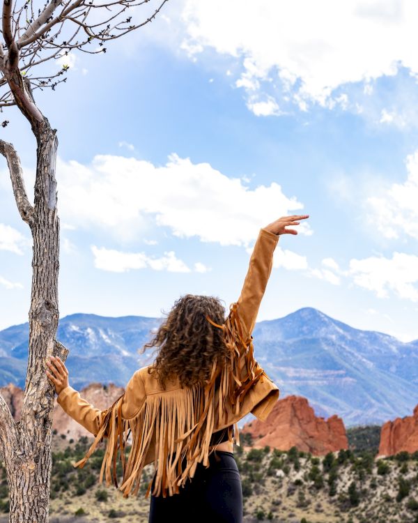 A person in a fringed jacket stands near a tree, raising their arm, with a scenic backdrop of mountains and a partly cloudy sky.