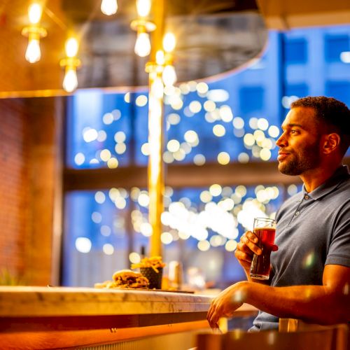 A man is holding a drink at a bar with warm lighting and a blurred background featuring string lights and a window with a cityscape.