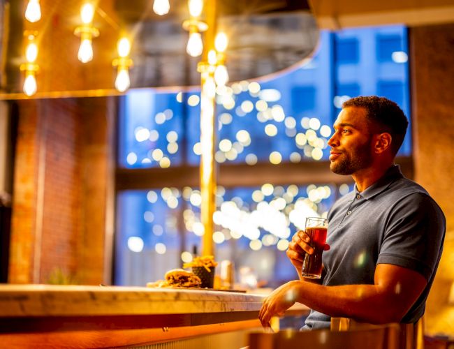 A man is holding a drink at a bar with warm lighting and a blurred background featuring string lights and a window with a cityscape.